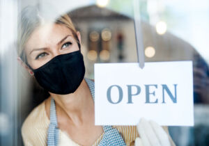 Coffee shop woman owner with face mask, open after lockdown quarantine. Shot through glass.