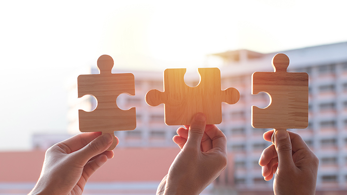 Hands are raised against a bright, white sky, each holding a piece of a wooden jigsaw puzzle.