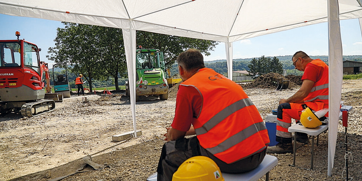 People wearing high‑visibility vests sit under a canopy on a construction site, with several construction vehicles in the background.