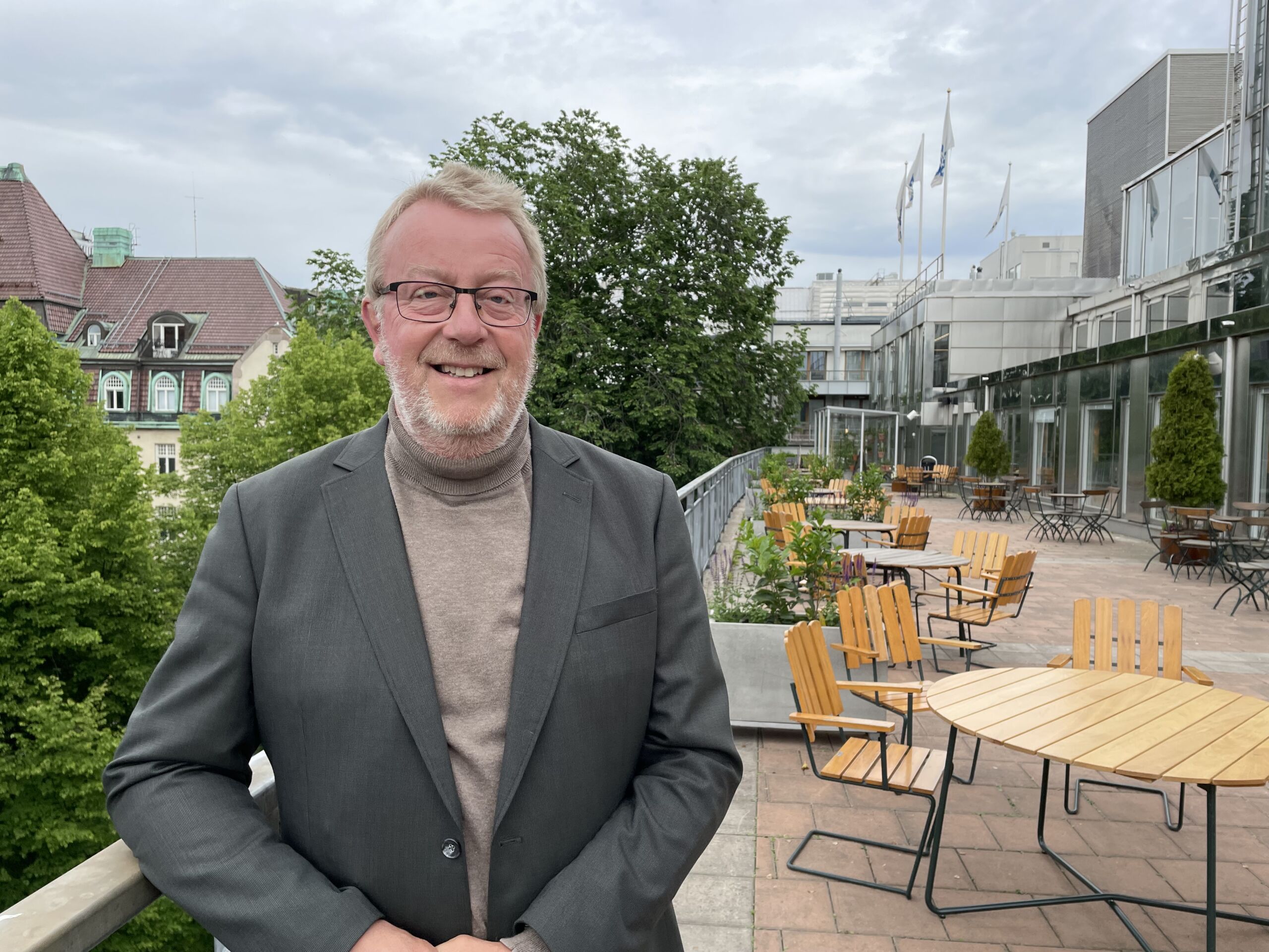 A man smiles while standing on an outdoor terrace with wooden tables; modern buildings and greenery appear in the background.